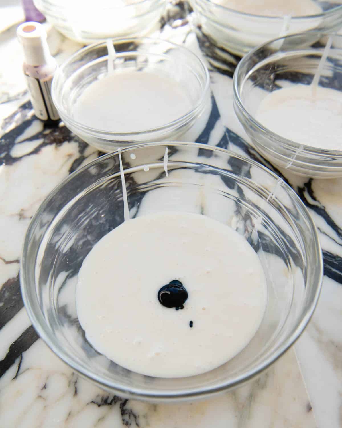 cut out sugar cookie frosting in a glass bowl with a drop of green food coloring in it before mixing with other bowls of white frosting and gel food coloring bottles in the background