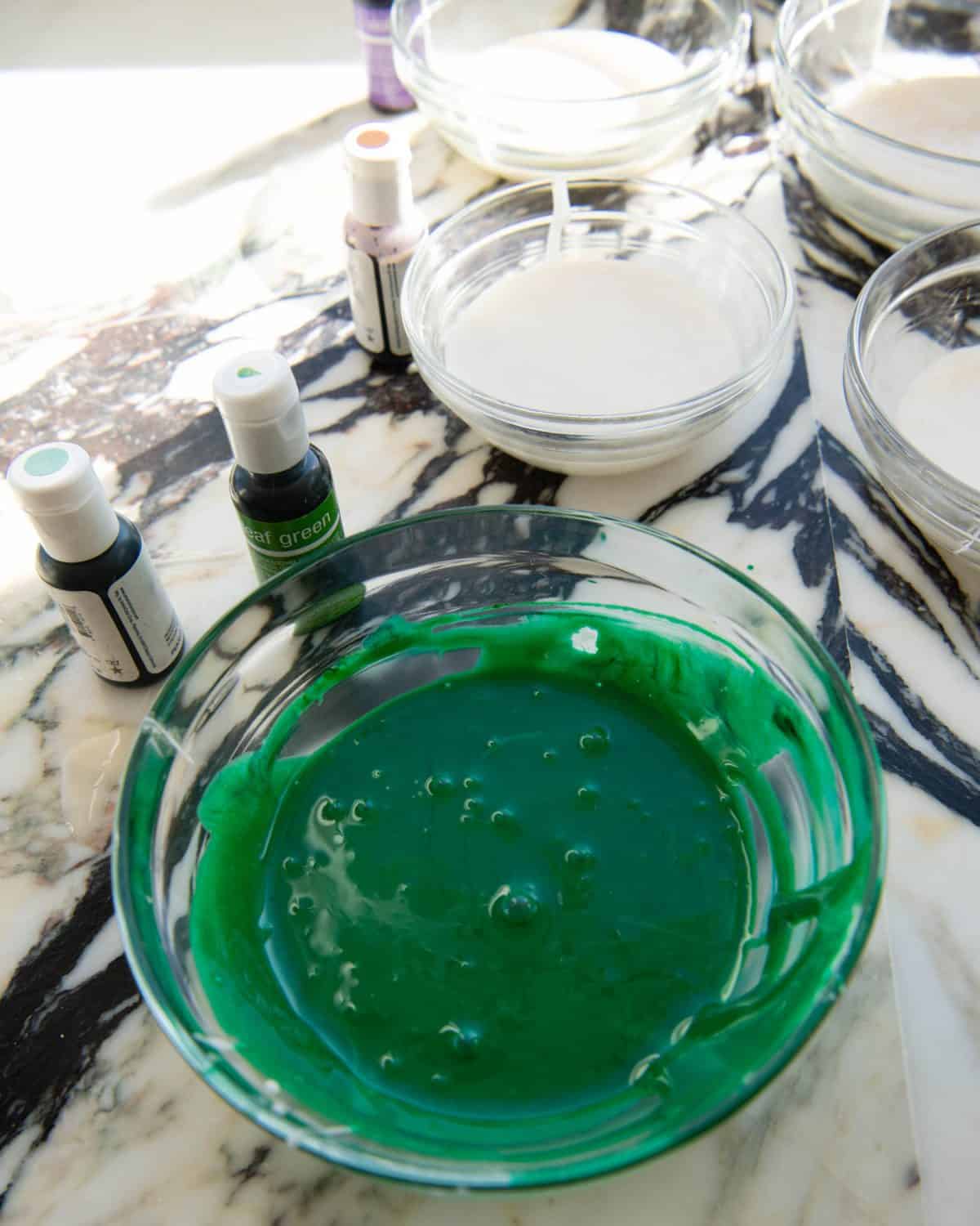 green cut out sugar cookie frosting in a glass bowl with other bowls of white frosting and gel food coloring bottles in the background