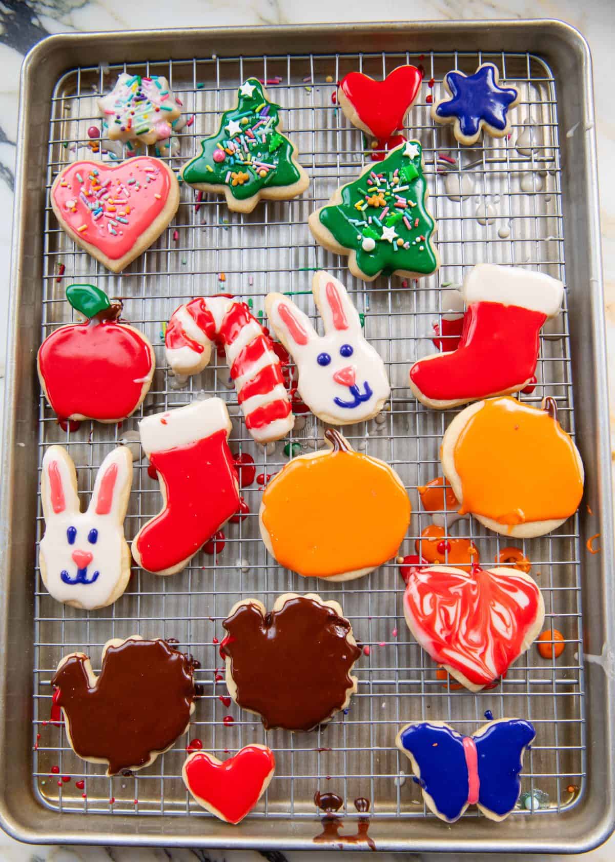frosted cut out sugar cookies in a metal baking sheet lined with a metal wire rack just after frosting in the shapes of hearts, Christmas trees, stockings, pumpkin, turkeys, bunnies and a butterfly.