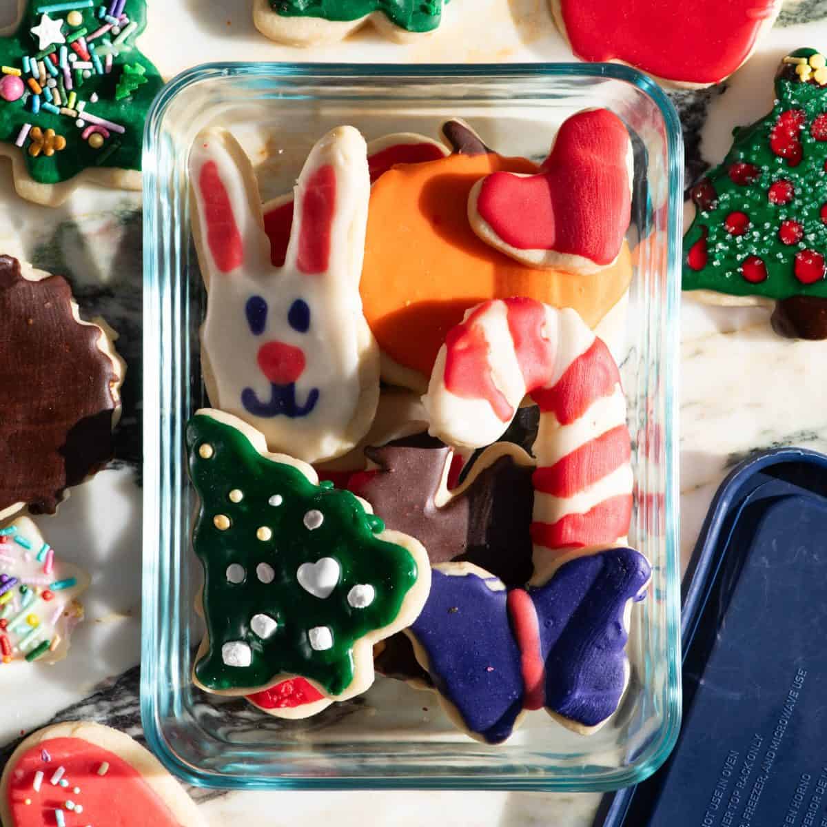 frosted cut out sugar cookies in a glass container to be stored in the shapes of a Christmas tree, butterfly, bunny, turkey, candy cane, pumpkin and hear