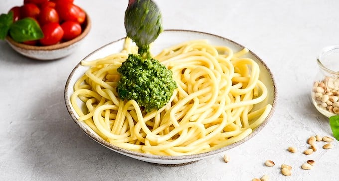 front view of a spoon putting homemade pesto sauce over a bowl of spaghetti noodles to make Pesto Pasta