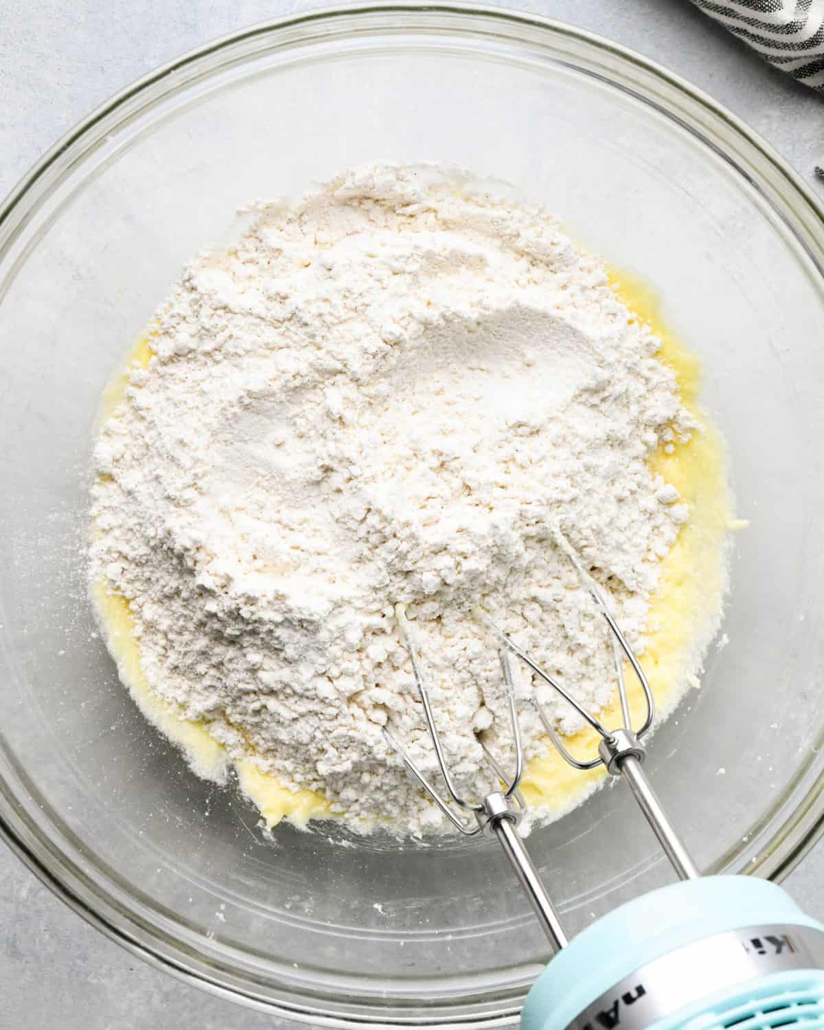 making snickerdoodles - flour mixture added to the wet mixture in a glass bowl before mixing. There's a small blue handheld mixer with two beaters in the bowl too