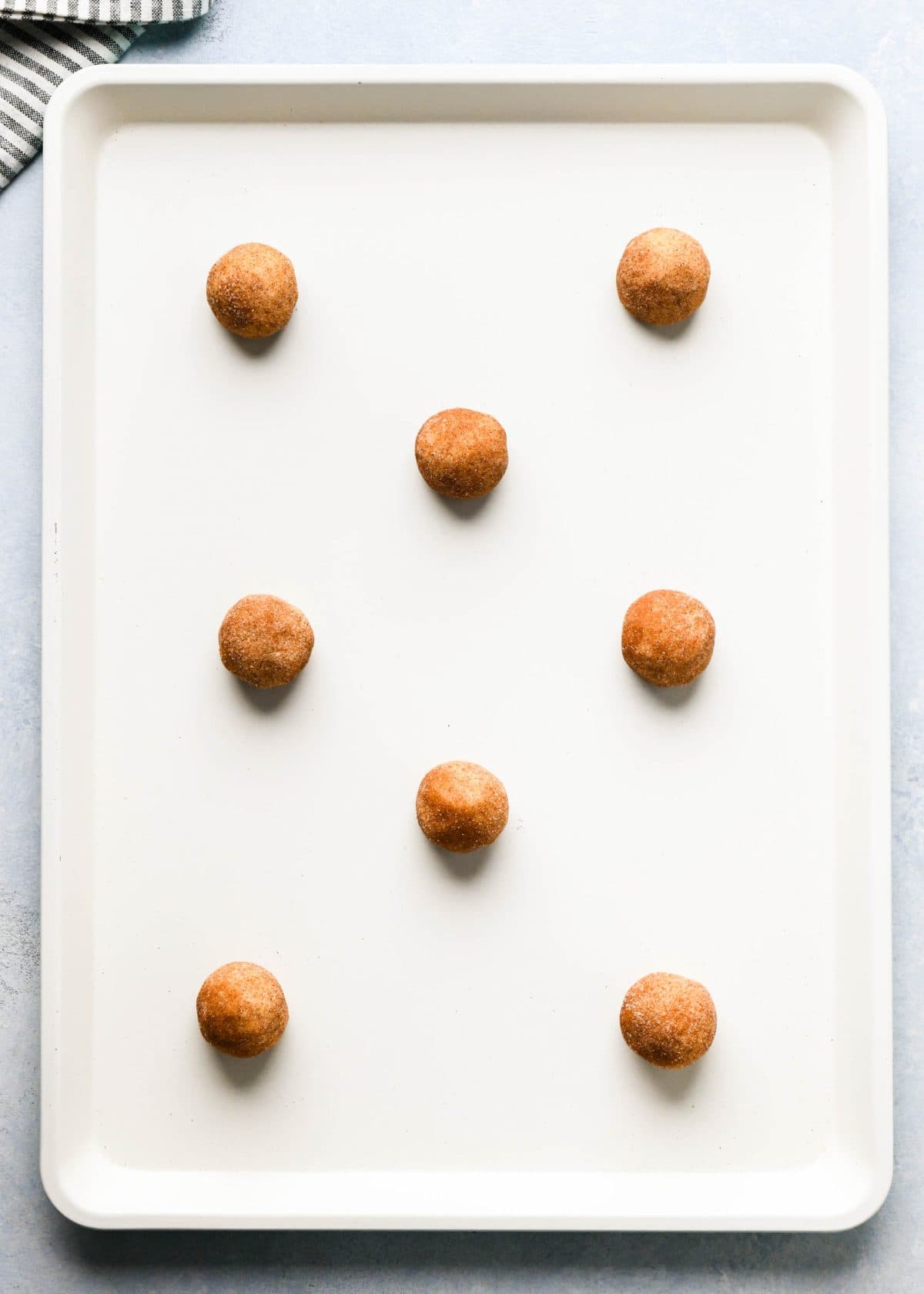 making snickerdoodles - 8 cookies rolled in cinnamon sugar on a white baking sheet lined with white parchment paper before baking