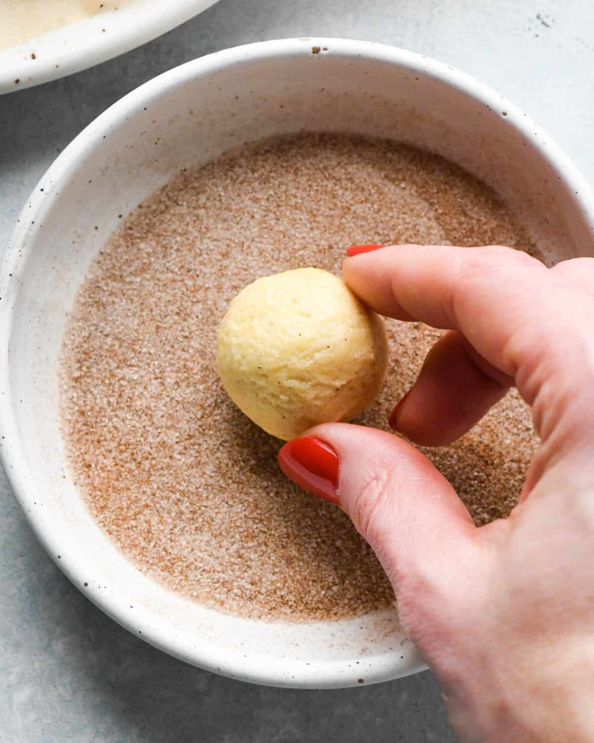 making snickerdoodles - a hand dipping a cookie dough ball into the bowl of cinnamon sugar