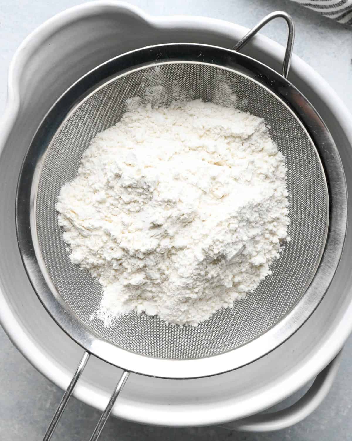 making snickerdoodles - sifting flour in a fine mesh metal strainer over a white ceramic mixing bowl