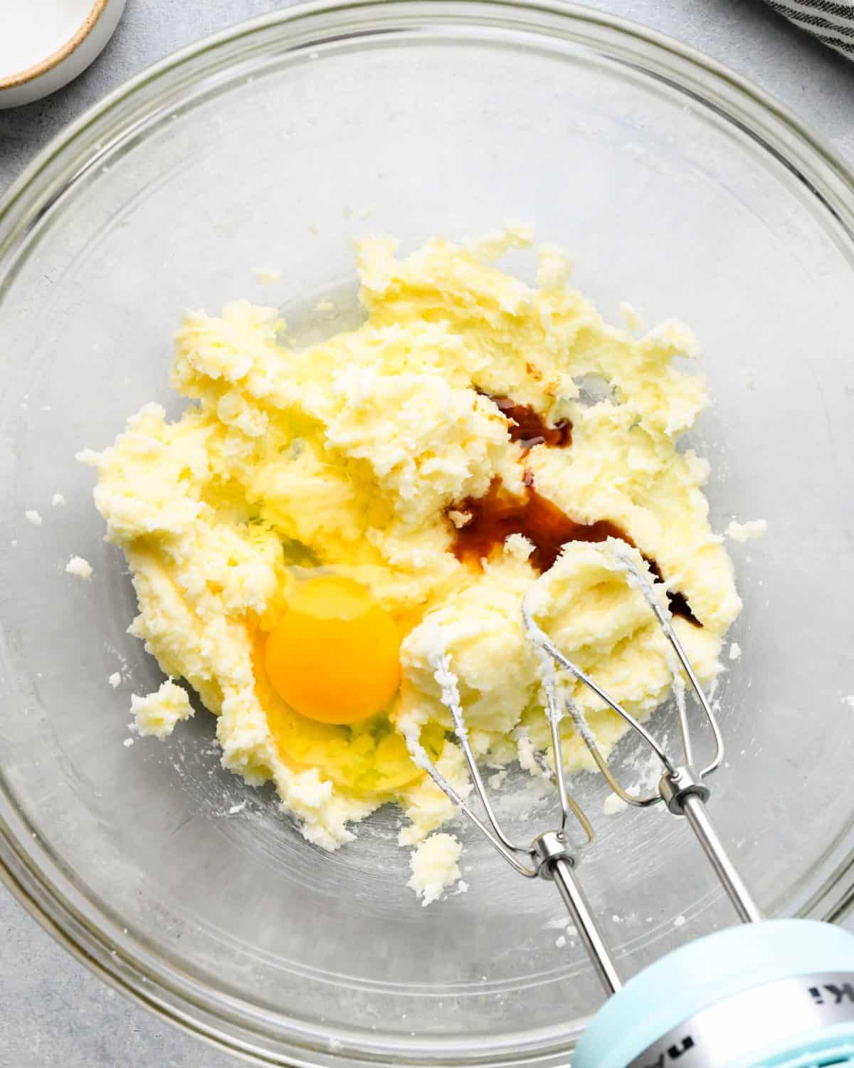 making snickerdoodles - egg and vanilla added to the beaten butter mixture in a glass mixing bowl with a small blue hand mixer with two beaters before mixing