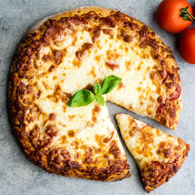 overhead view of a round pizza with a Zucchini Pizza Crust topped with cheese with one triangular slice cut out of it sitting in it's place and a sprig of basil in the center of the zucchini pizza. 