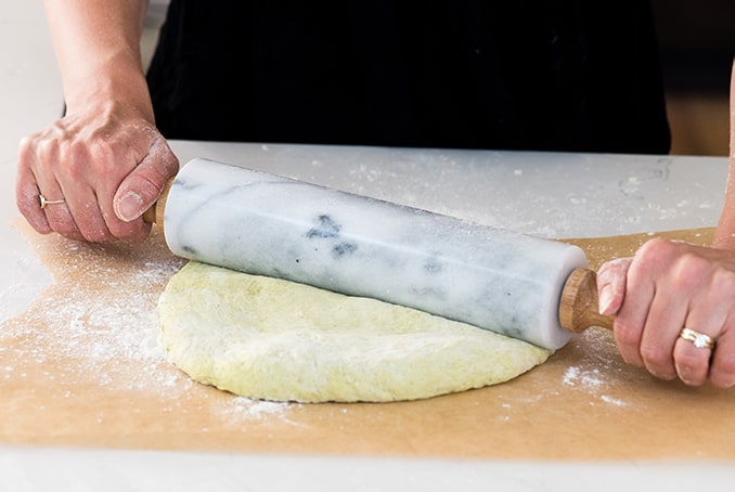 front view of hands rolling out a Zucchini Pizza Crust on a piece of brown parchment paper with a marble rolling pin