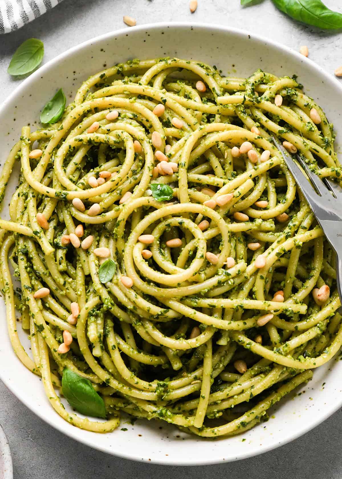 close up photo of pesto pasta in a ceramic bowl with a silver fork, garnished with basil and toasted pine nuts on a gray surface 
