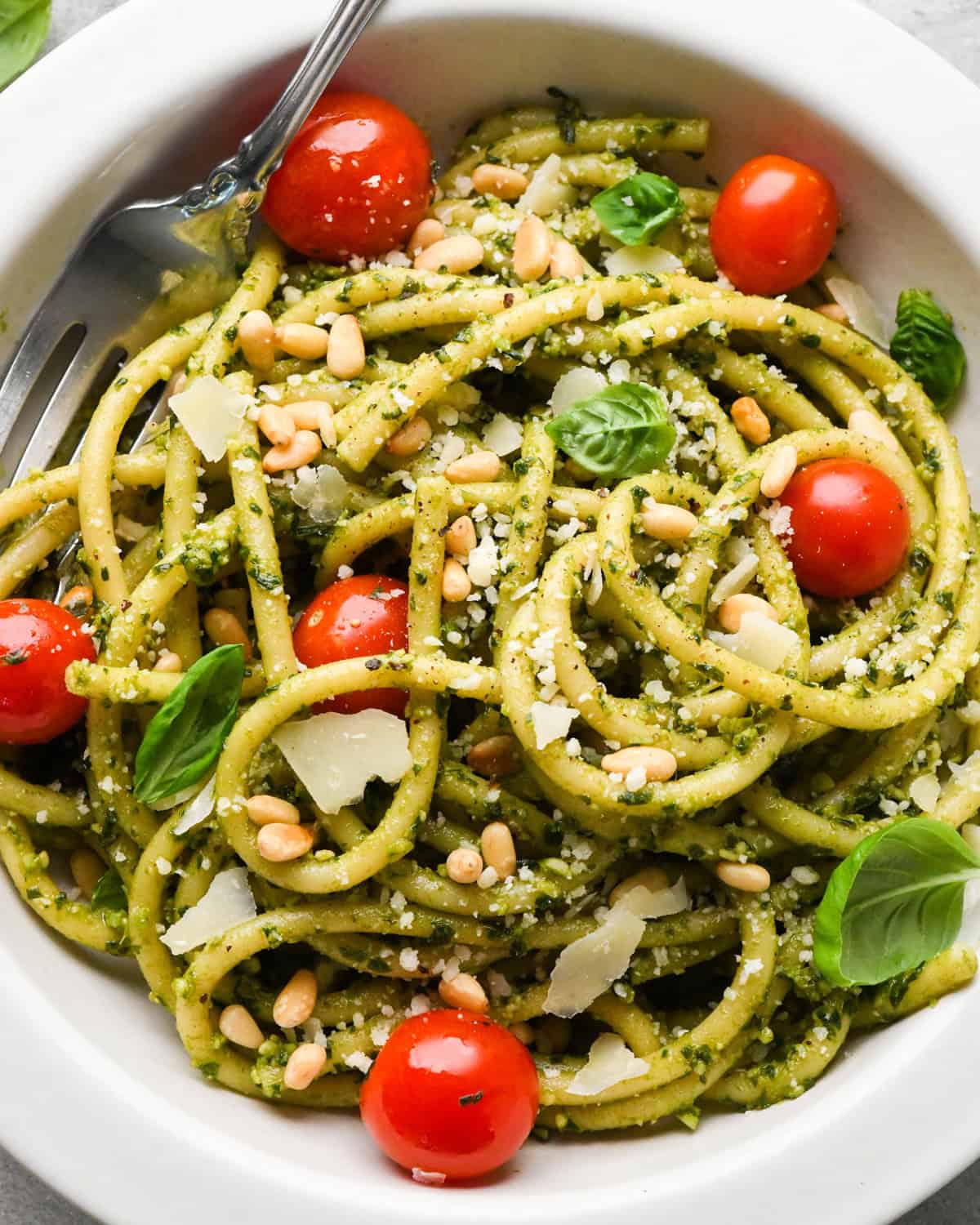 pesto pasta in a white bowl with cherry tomatoes and a silver fork, garnished with toasted pine nuts, parmesan cheese and fresh basil 