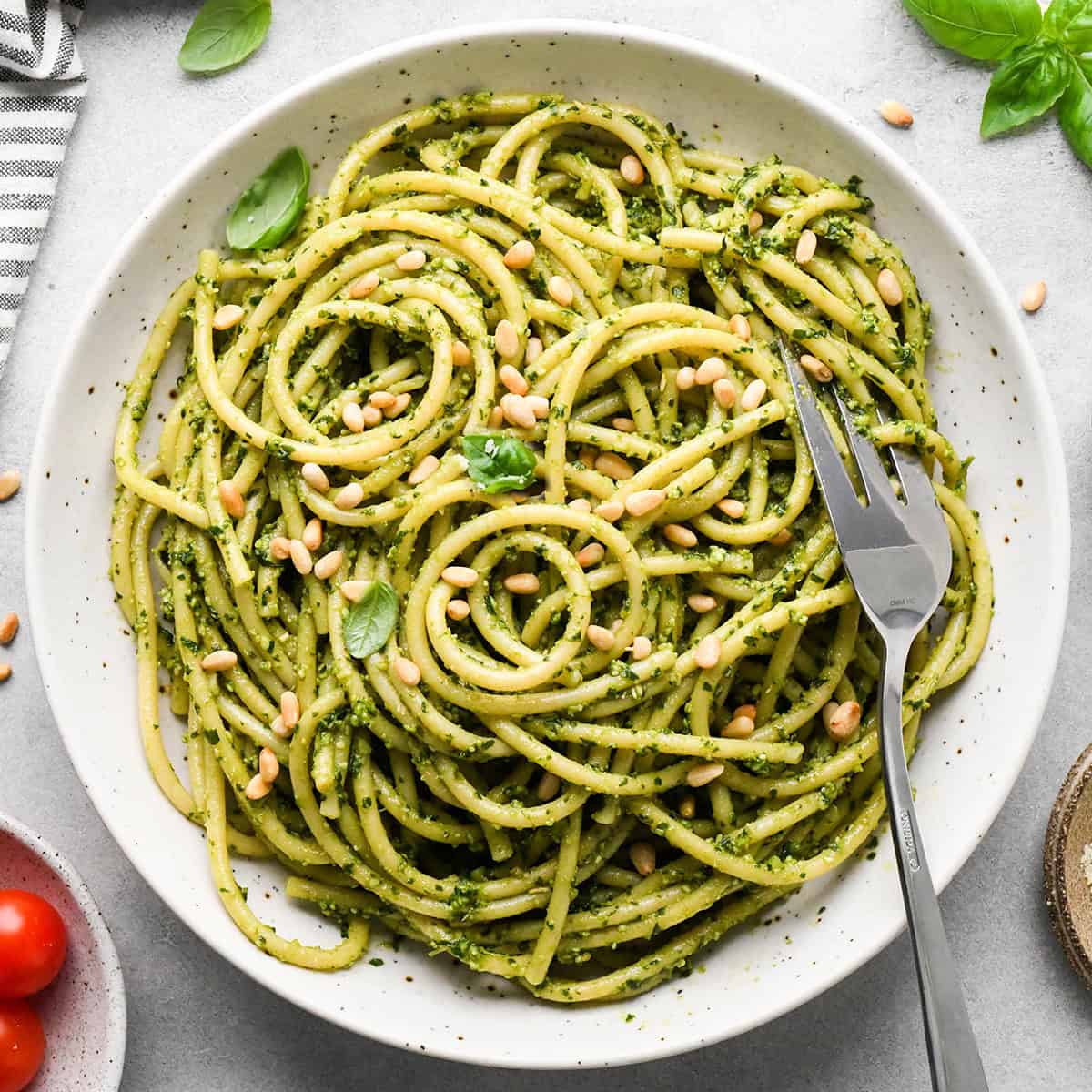 pesto pasta in a ceramic bowl with a silver fork, garnished with toasted pine nuts and fresh basil on a gray surface surrounded by basil, pine nuts, tomatoes and a striped towel. 