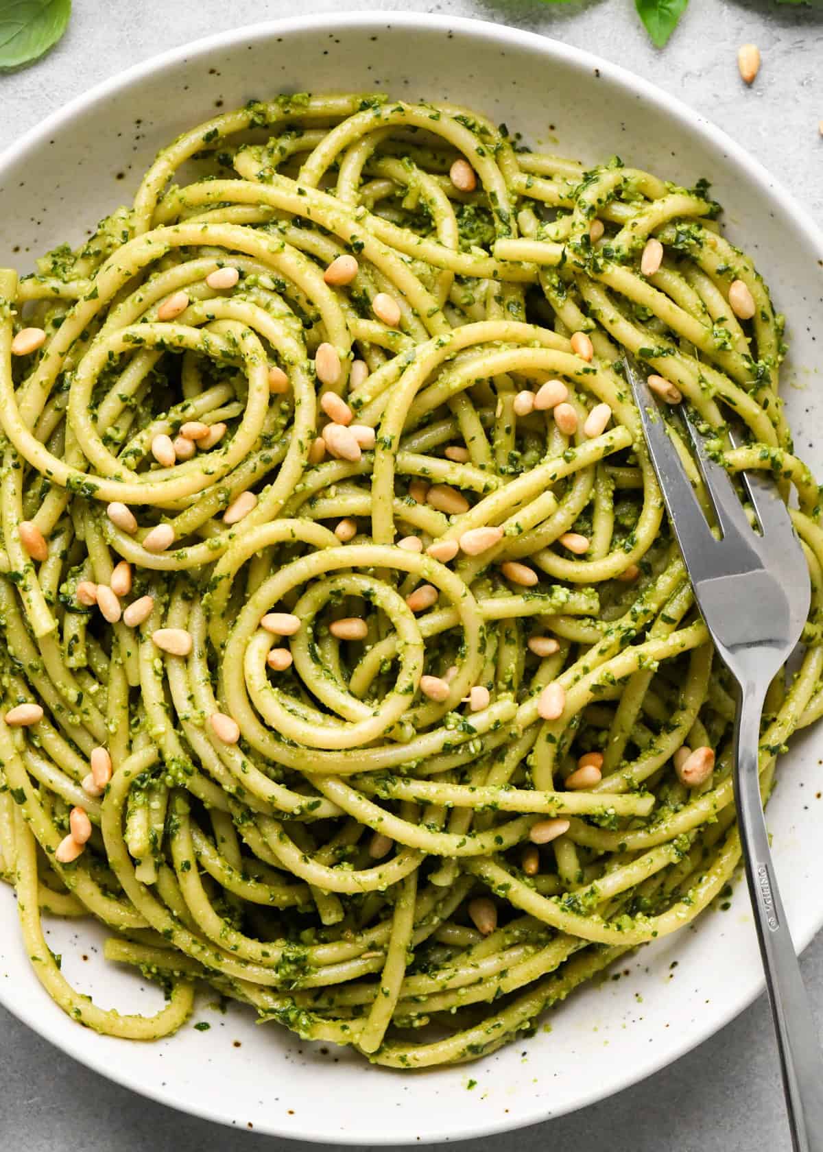 close up photo of pesto pasta in a ceramic bowl with a silver fork, garnished with toasted pine nuts on a gray surface 