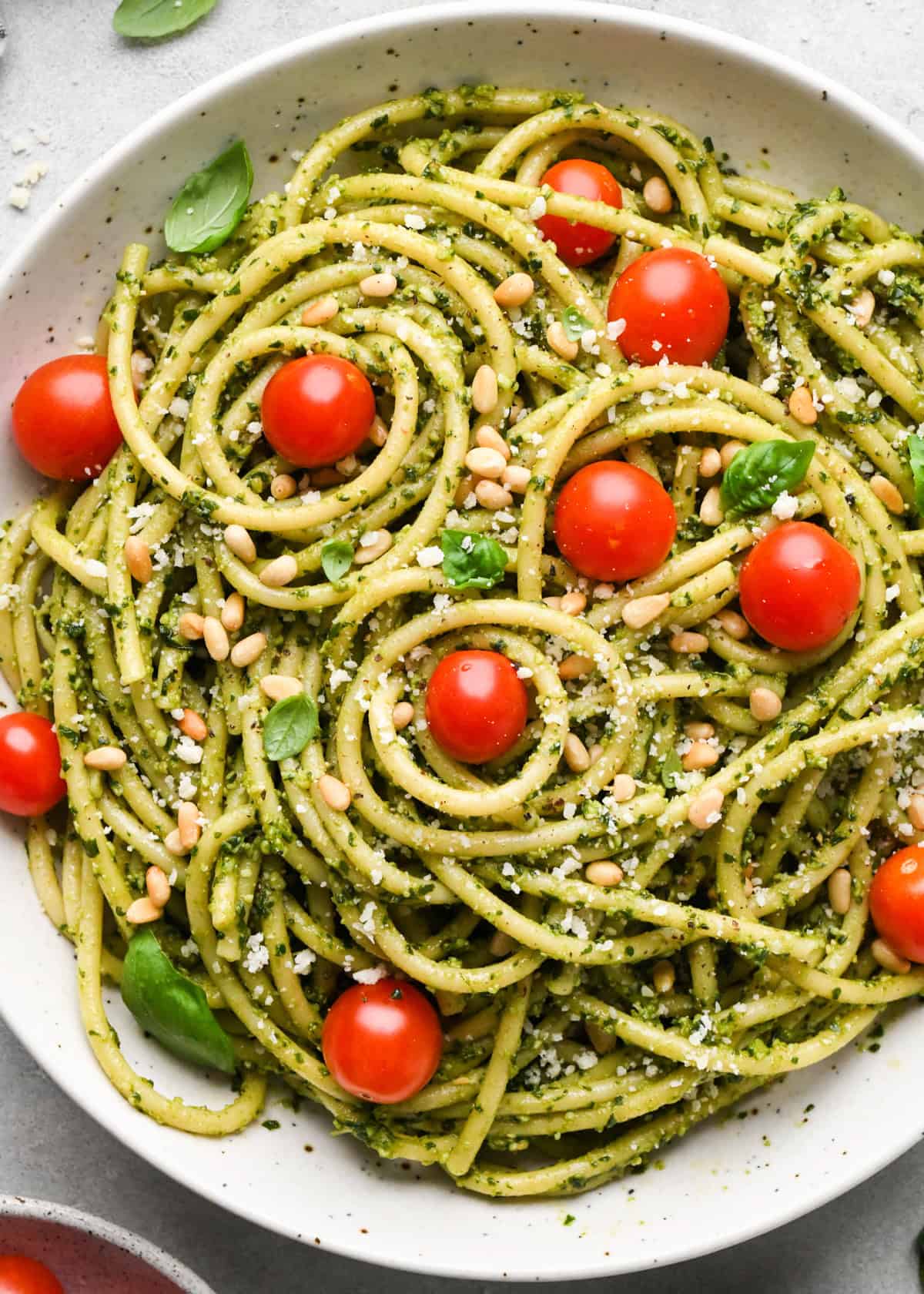 pesto pasta in a ceramic bowl with cherry tomatoes and a silver fork, garnished with toasted pine nuts, parmesan cheese and fresh basil on a gray surface 