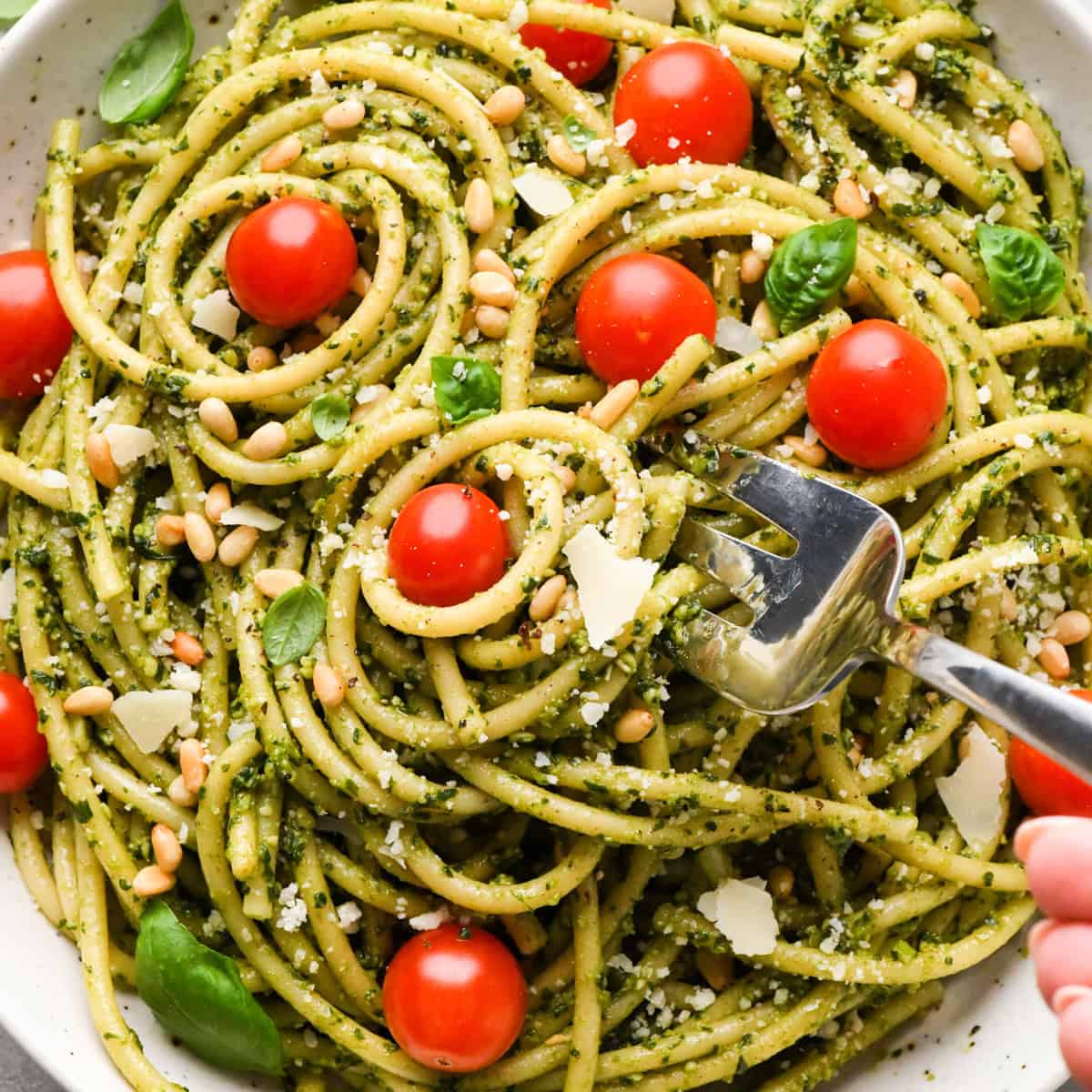 a fork taking a bite of pesto pasta in a ceramic bowl with cherry tomatoes and a silver fork, garnished with toasted pine nuts, parmesan cheese and fresh basil on a gray surface surrounded by basil, pine nuts, tomatoes and a striped towel. 