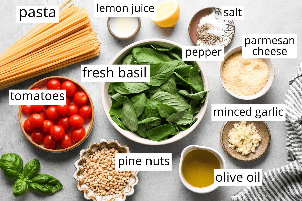 overhead photo of the ingredients in this pesto pasta recipe labeled and in bowls like fresh basil, tomatoes, pasta, minced garlic, olive oil, pine nuts, parmesan cheese and lemon juice. 
