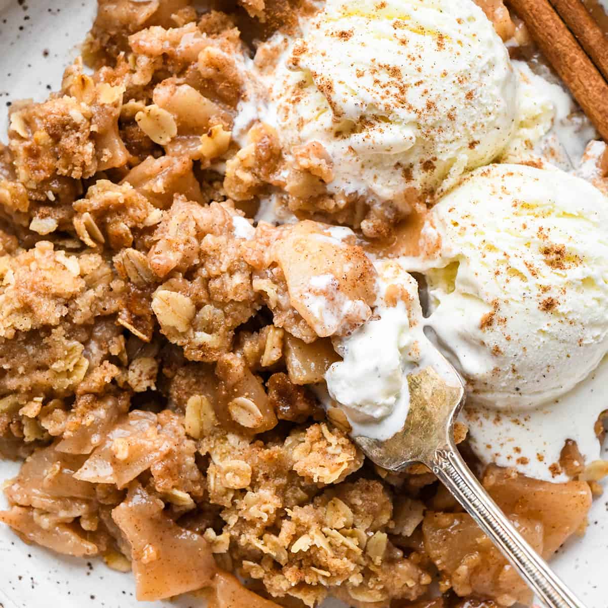 up close photo of a serving of apple crisp on a plate with vanilla ice cream and a fork