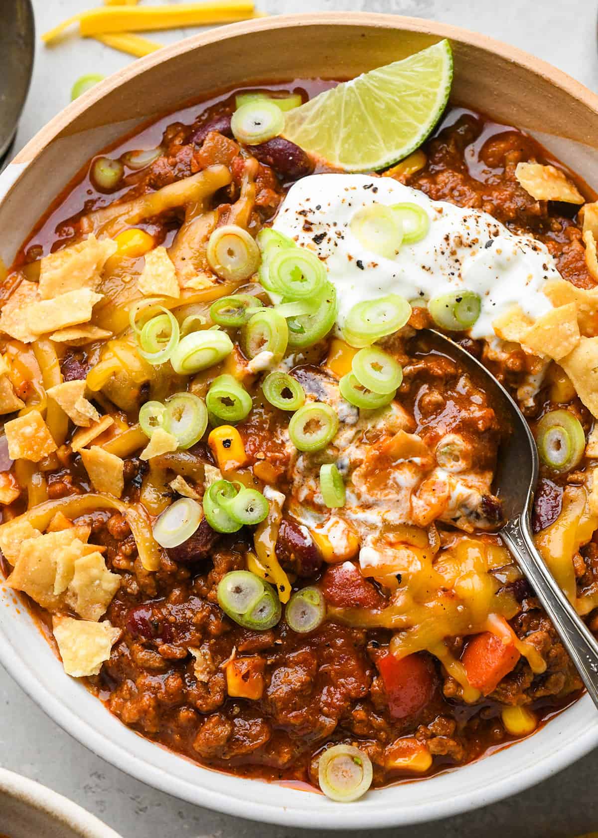homemade chili in a bowl with a spoon topped with sour cream, onions and cheese