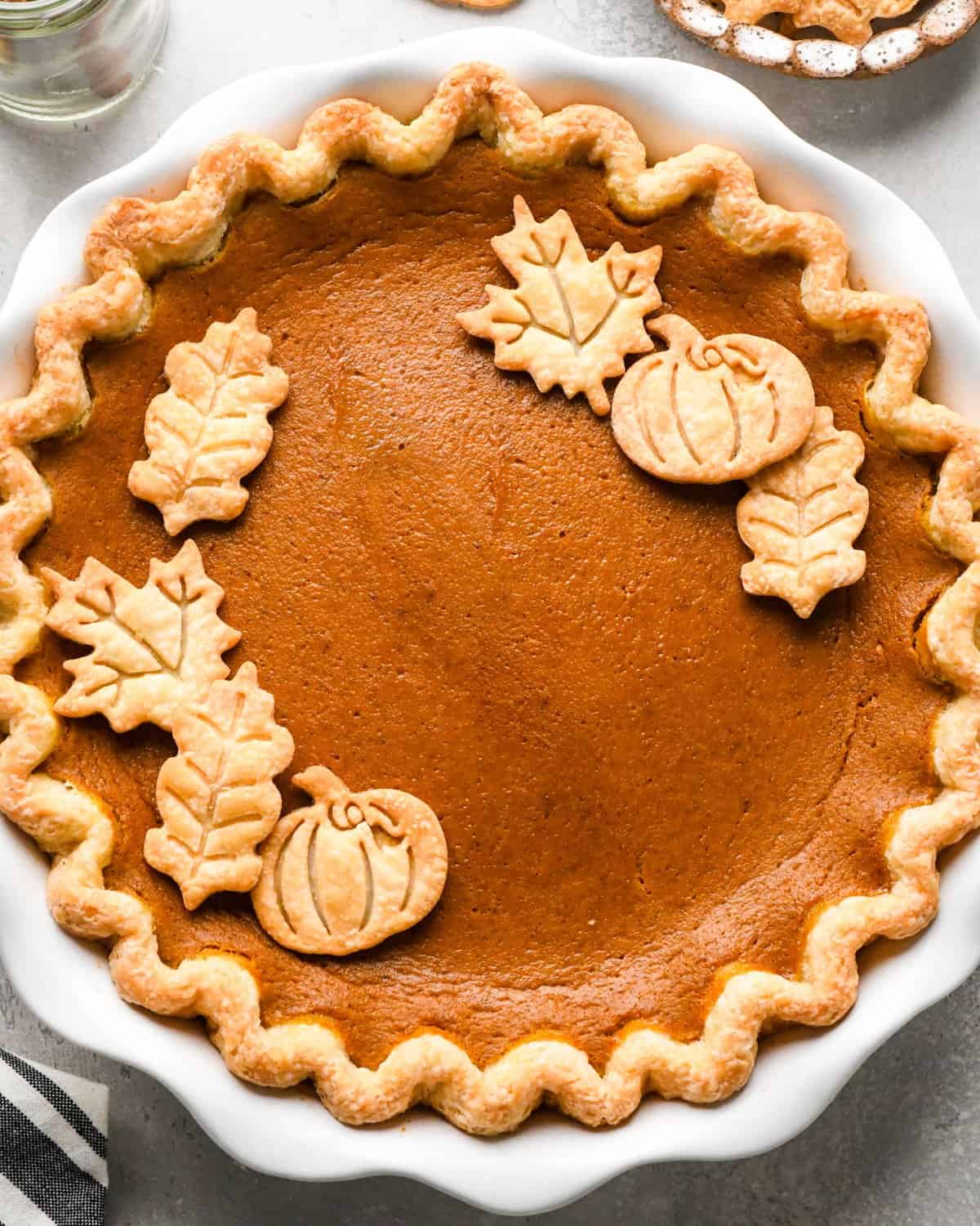 Pumpkin pie in a white pie dish with decorative pumpkin and leaf-shaped crust toppings, set on a gray surface with a striped towel nearby.