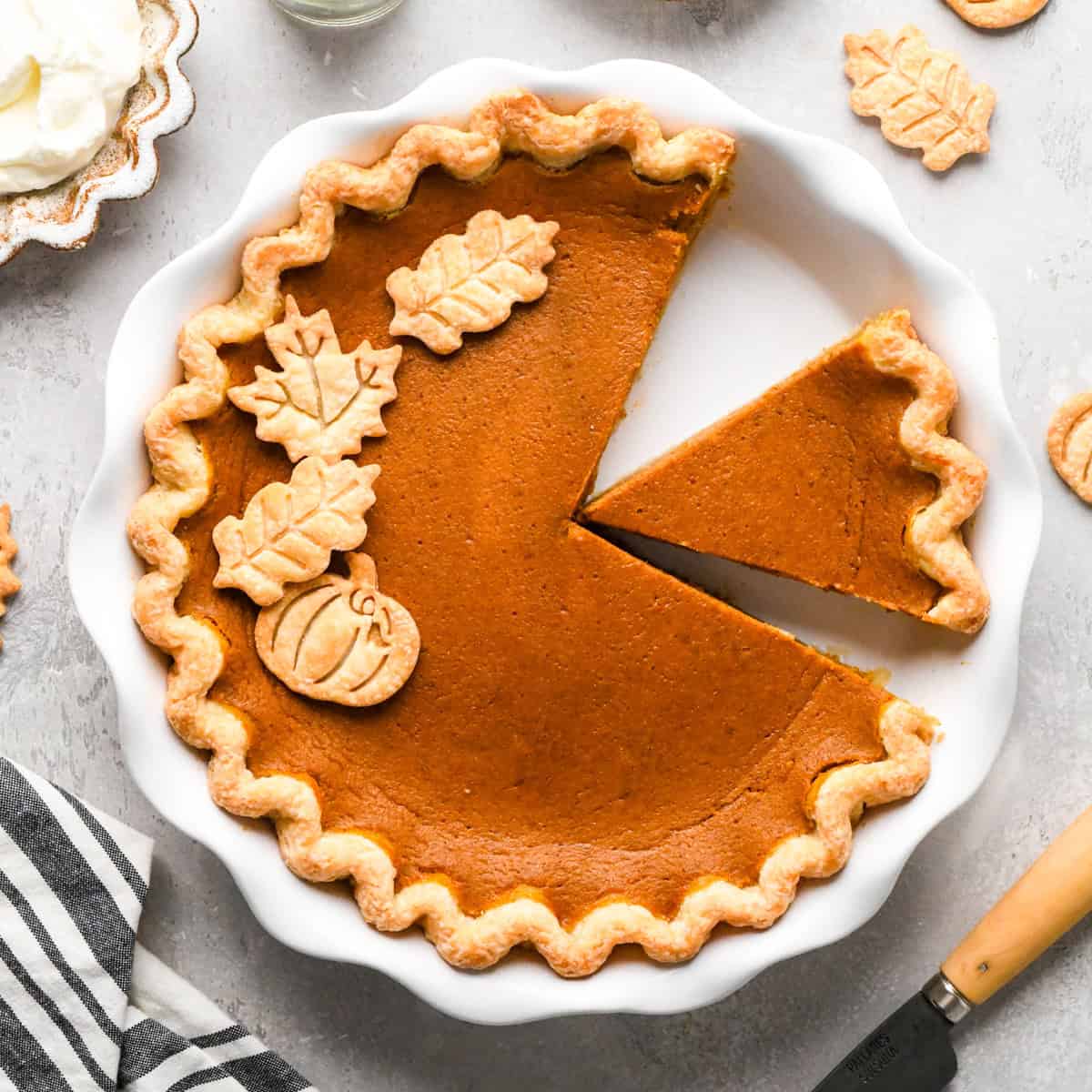 Pumpkin pie in a white pie dish with one slice cut out with decorative pumpkin and leaf-shaped crust toppings, set on a gray surface with a striped towel and whipped cream nearby. 