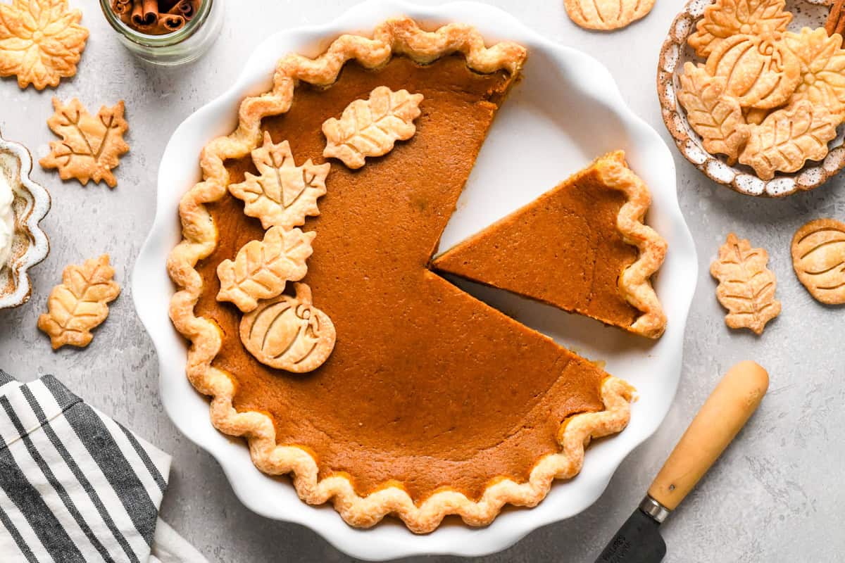 Pumpkin pie in a white pie dish with one slice cut out with decorative pumpkin and leaf-shaped crust toppings, set on a gray surface with a striped towel and whipped cream nearby. 
