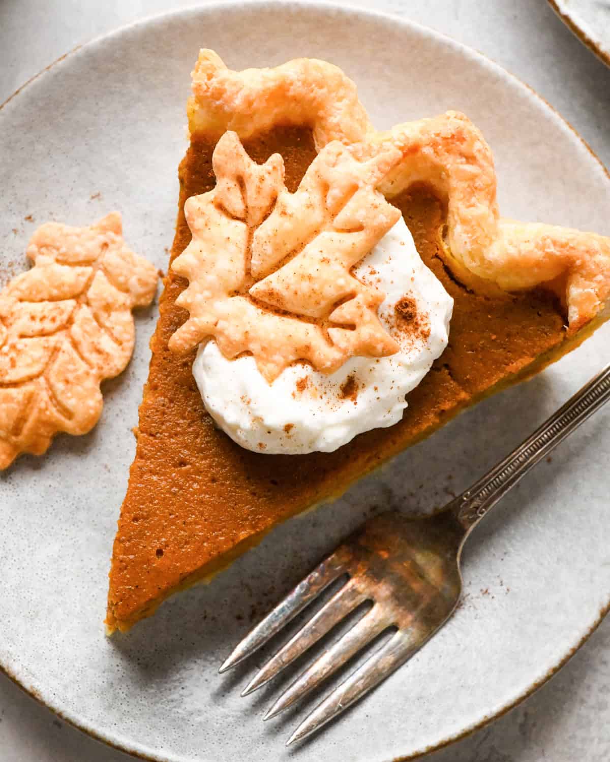 A slice of pumpkin pie topped with a dollop of whipped cream and a pie crust leaf with a fork next to it on a gray plate.