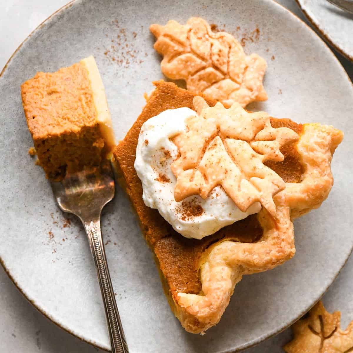 A slice of pumpkin pie topped with a dollop of whipped cream and a pie crust leaf with a fork with a bite on it taken out of it on a gray plate.