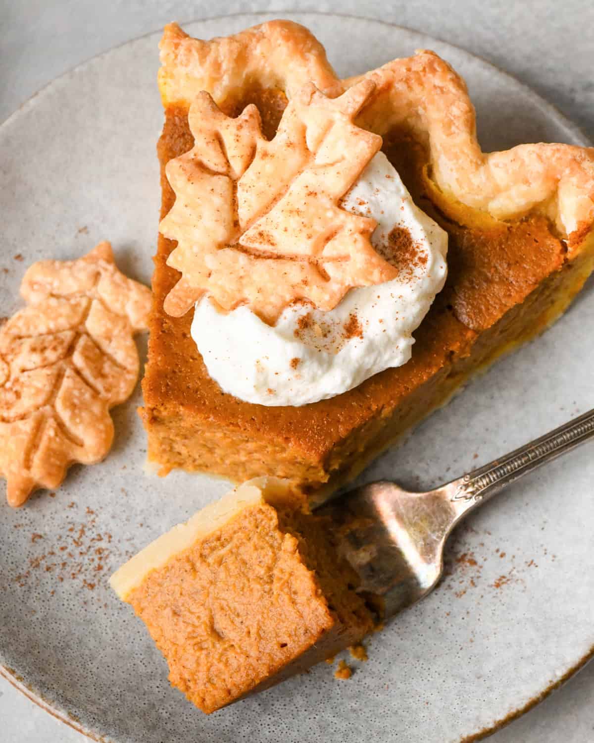 A slice of pumpkin pie topped with a dollop of whipped cream and a pie crust leaf with a fork with a bite on it taken out of it on a gray plate.