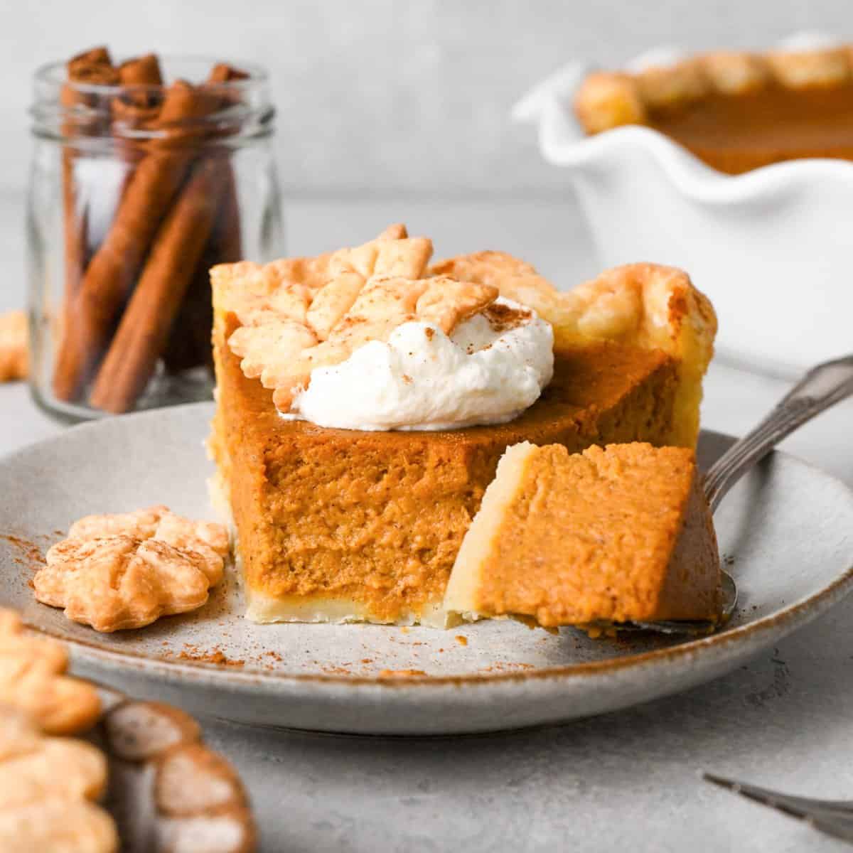 A slice of pumpkin pie topped with a dollop of whipped cream and a pie crust leaf with a fork with a bite on it taken out of it on a gray plate. There are cinnamon sticks and a while pumpkin pie in the background