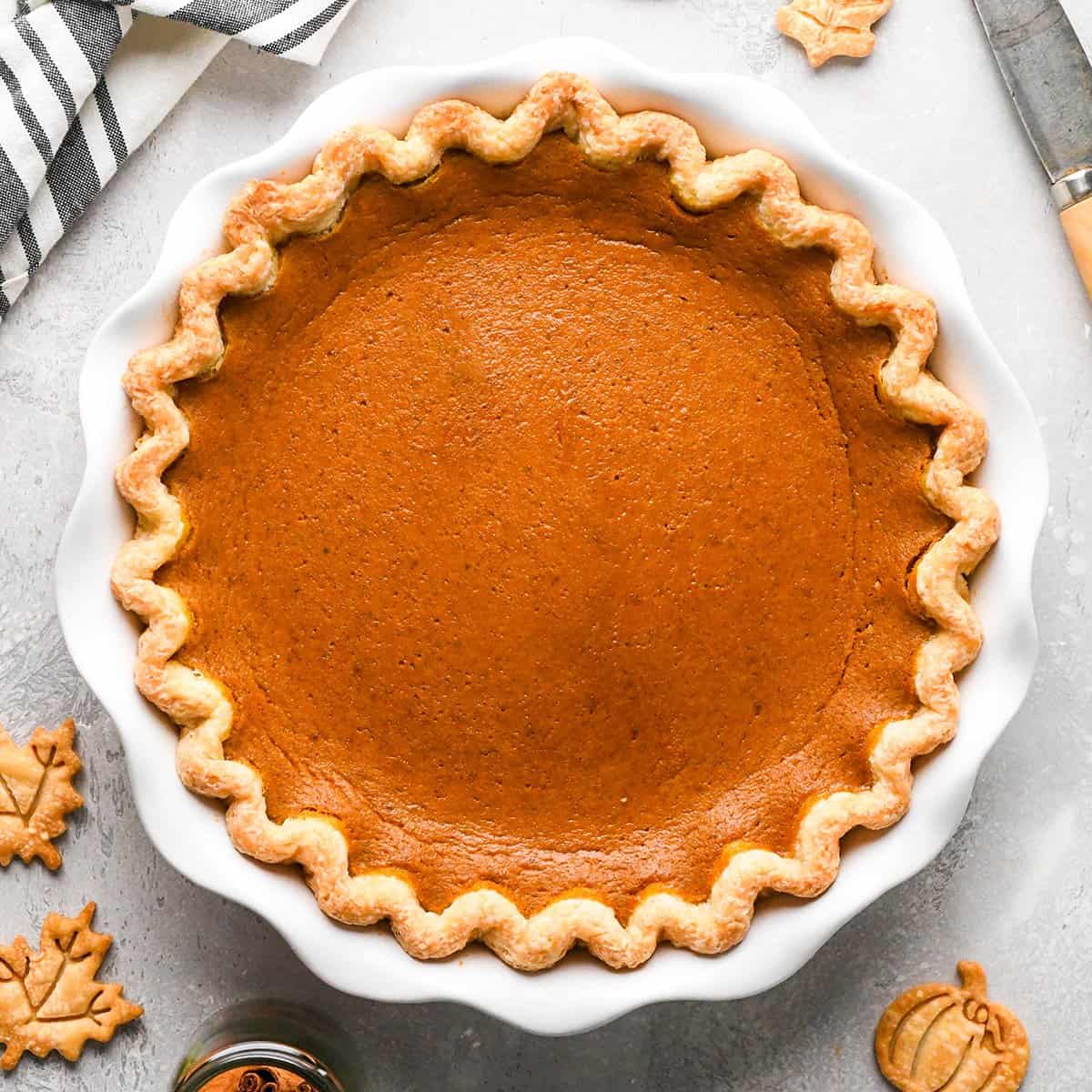 Pumpkin pie in a white pie dish set on a gray surface with a striped towel and pumpkin and leaf pie shapes nearby.