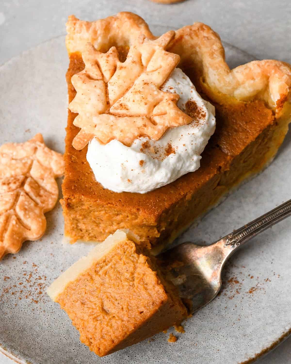 A slice of pumpkin pie topped with a dollop of whipped cream and a pie crust leaf with a fork with a bite on it taken out of it on a gray plate.