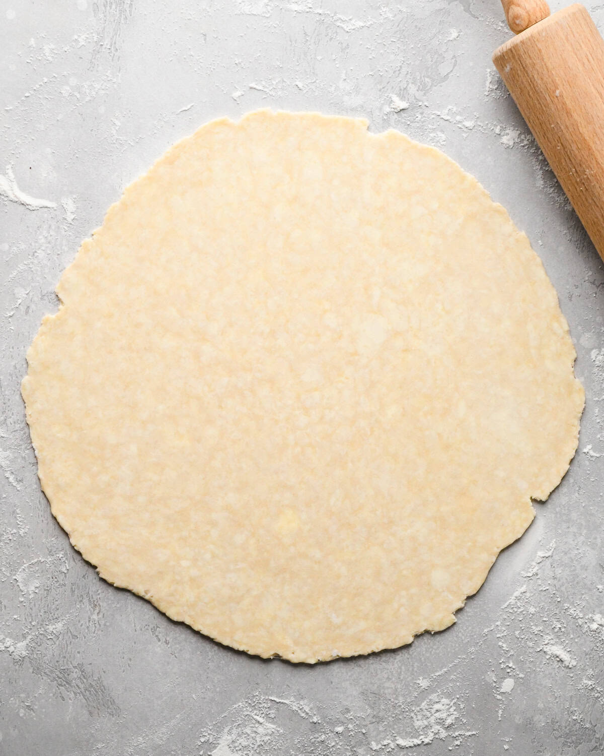 pie crust dough rolled out on a gray surface with flour and a wooden rolling pin