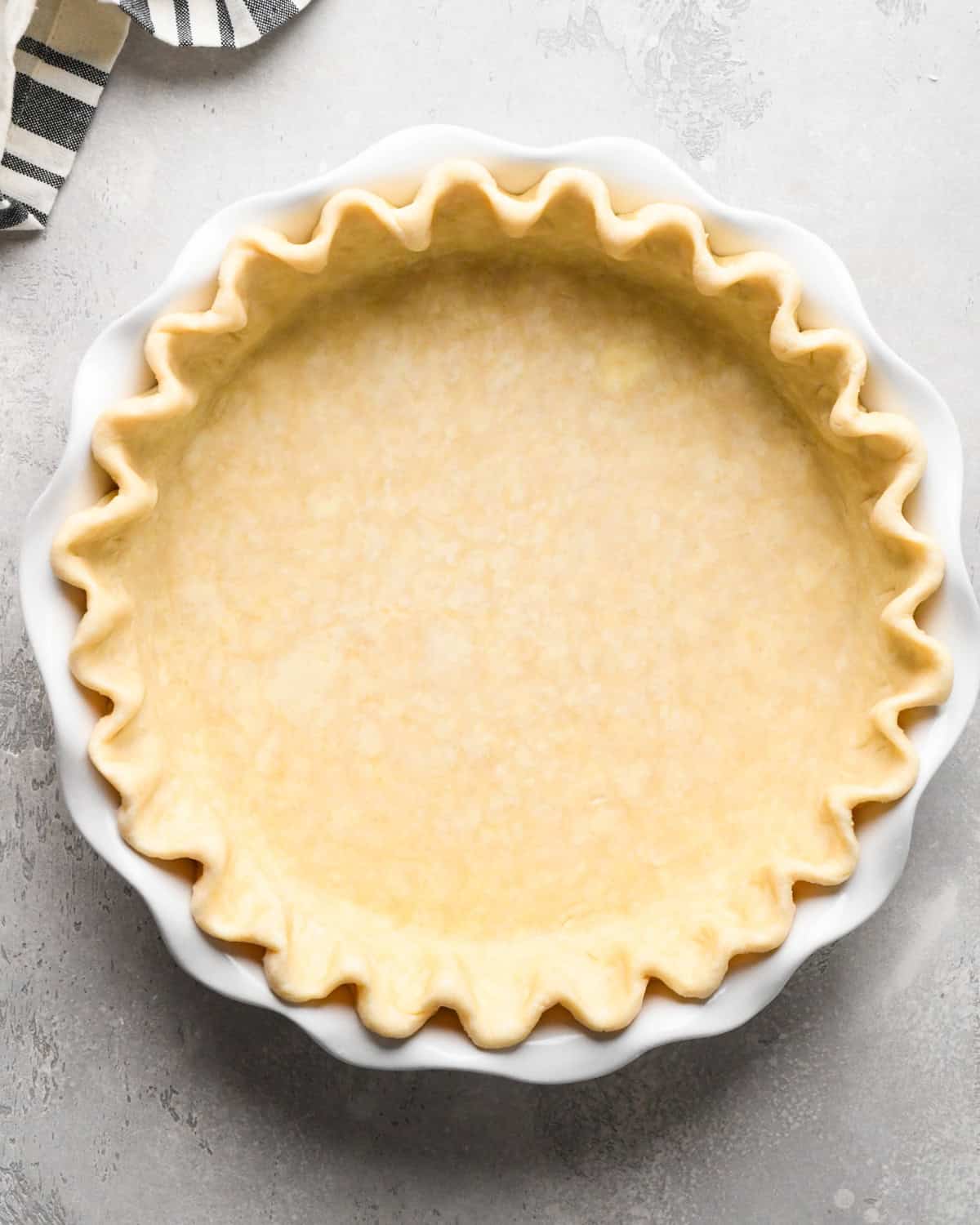 pie crust dough formed into a pie crust with fluted edges in a white ceramic pie dish on a gray surface to make pumpkin pie