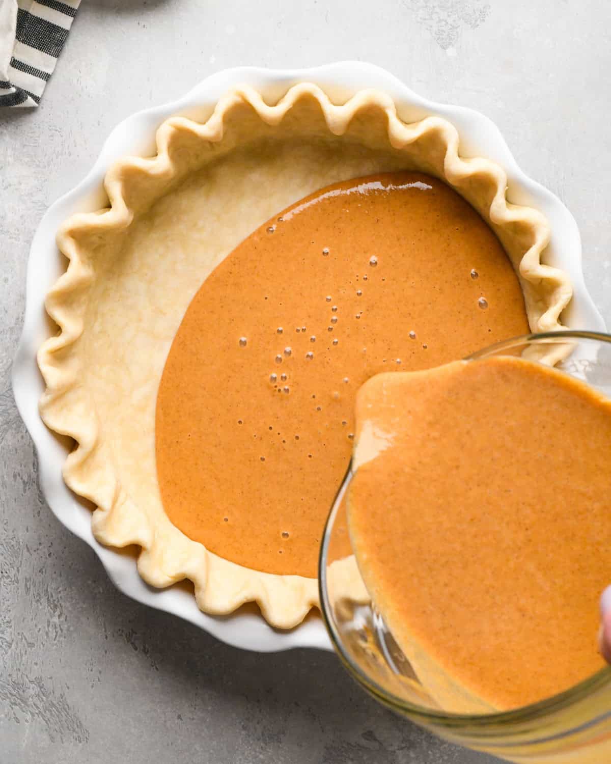 pumpkin pie filling being poured into the fluted pie crust in a white pie dish on a gray surface with a striped towel in the top left corner. 