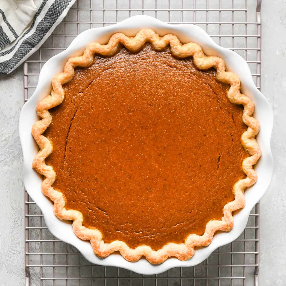 Pumpkin pie in a white pie dish on a wire cooling rack, set on a gray surface with a striped towel nearby.