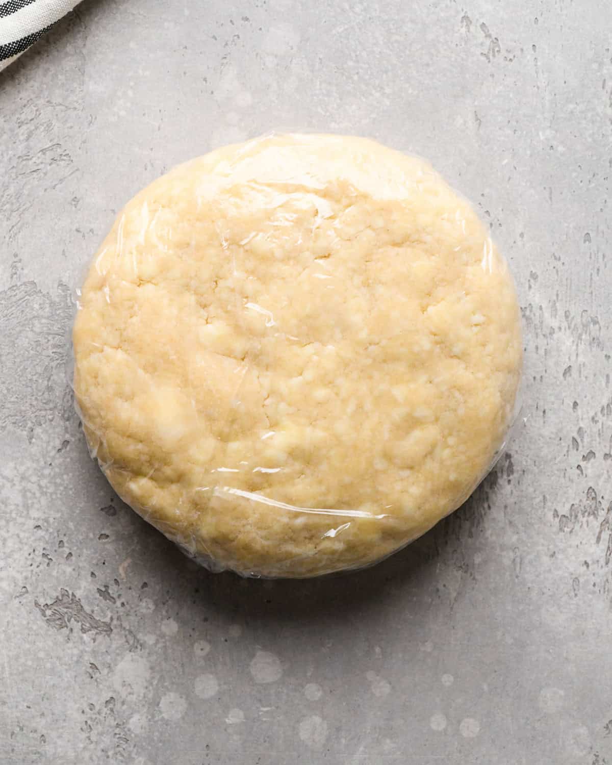 a round disc of pie crust dough wrapped in plastic wrap on a gray surface