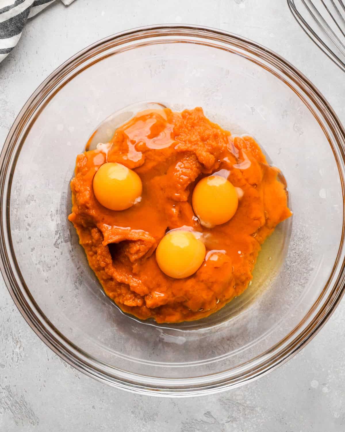 eggs and pumpkin puree in a glass bowl before whisking on a gray surface with a striped towel and whisk nearby making pumpkin pie