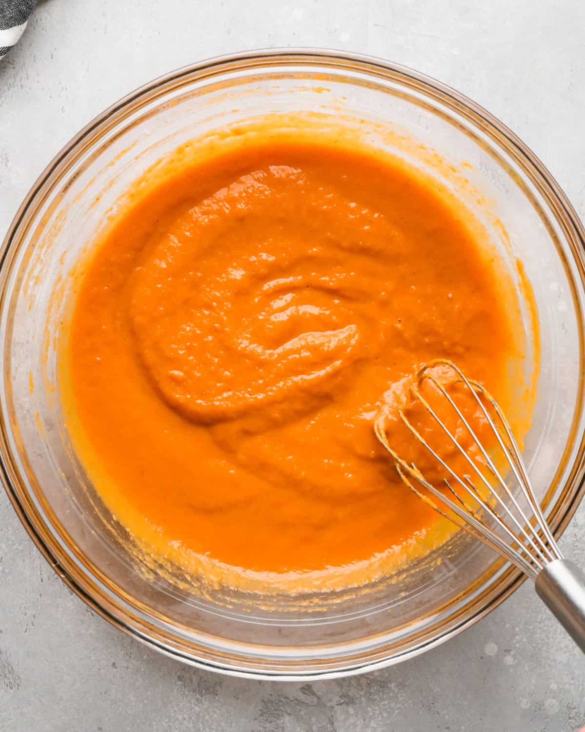 eggs and pumpkin puree whisked together in a glass bowl with a whisk on a gray surface with a striped towel in the top left corner