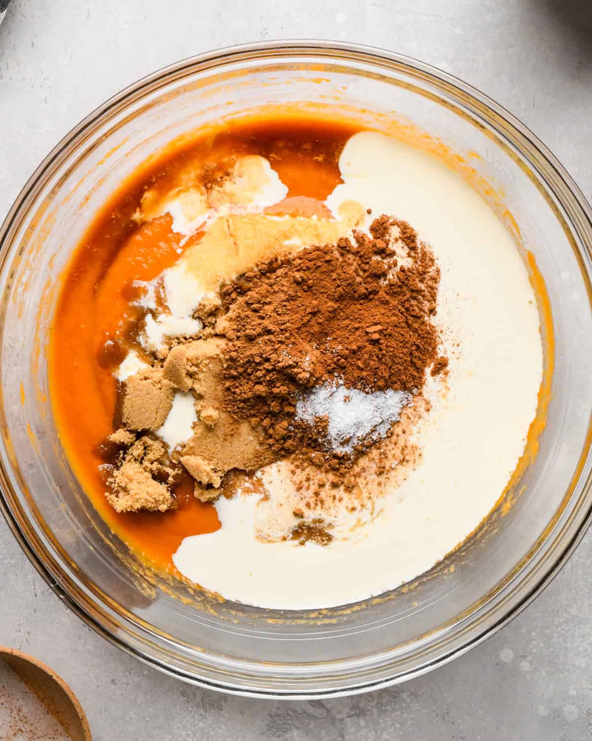 heavy cream, cinnamon, pumpkin pie spice, brown sugar and vanilla added to the pumpkin puree in a glass bowl before mixing on a gray surface making pumpkin pie