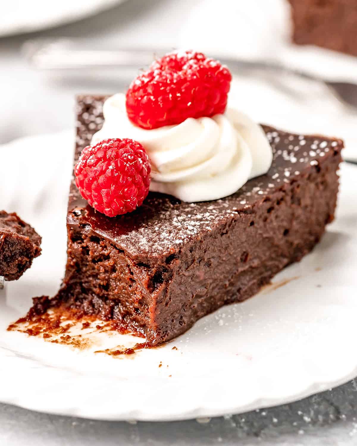 a slice of Flourless Chocolate Cake on a plate with a bite taken out of it, topped with whipped cream, powdered sugar and raspberries