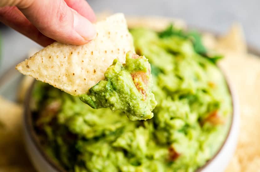 a hand holding a tortilla chip with guacamole on it over a bowl of guacamole surrounded by chips