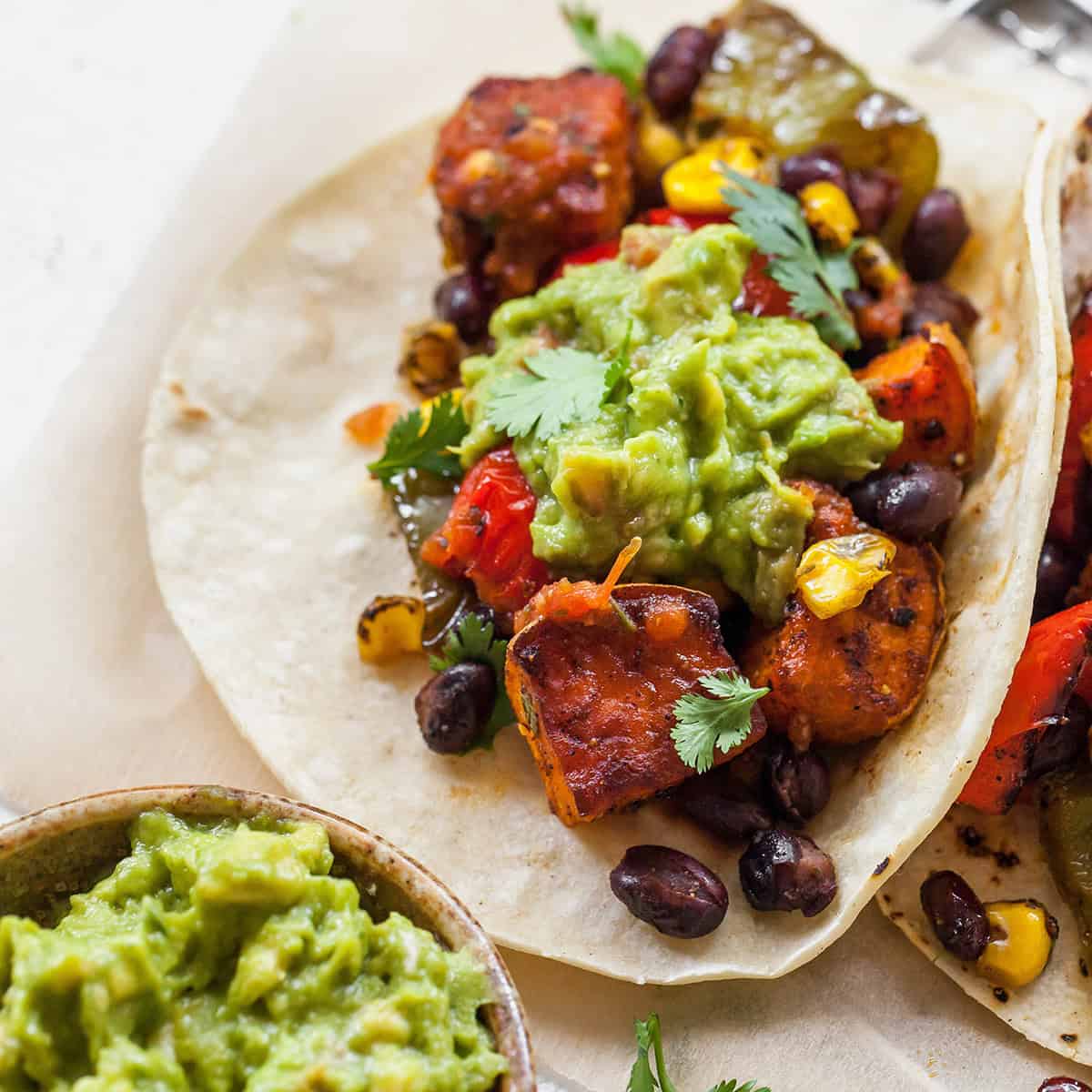 easy homemade guacamole recipe on a sweet potato taco with black beans, there's a bowl of guacamole in the bottom left corner 