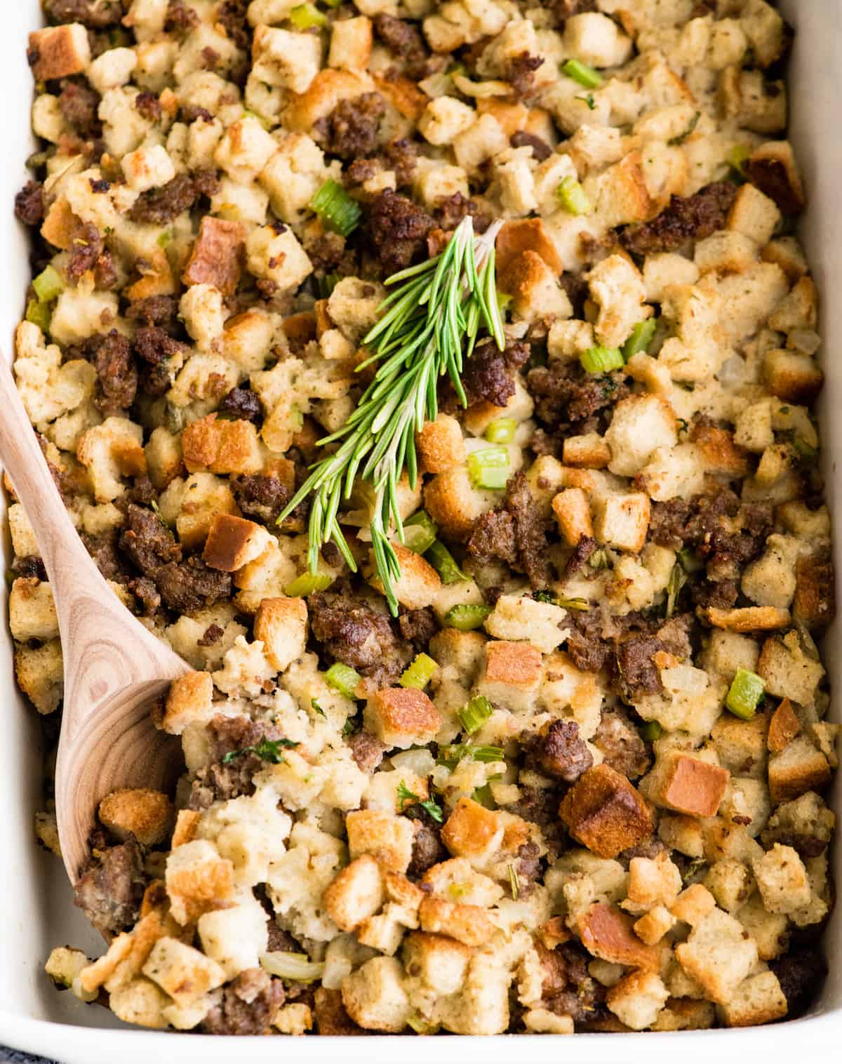 Overhead view of a baking dish of sausage stuffing with fresh rosemary on top and a spoon taking a scoop