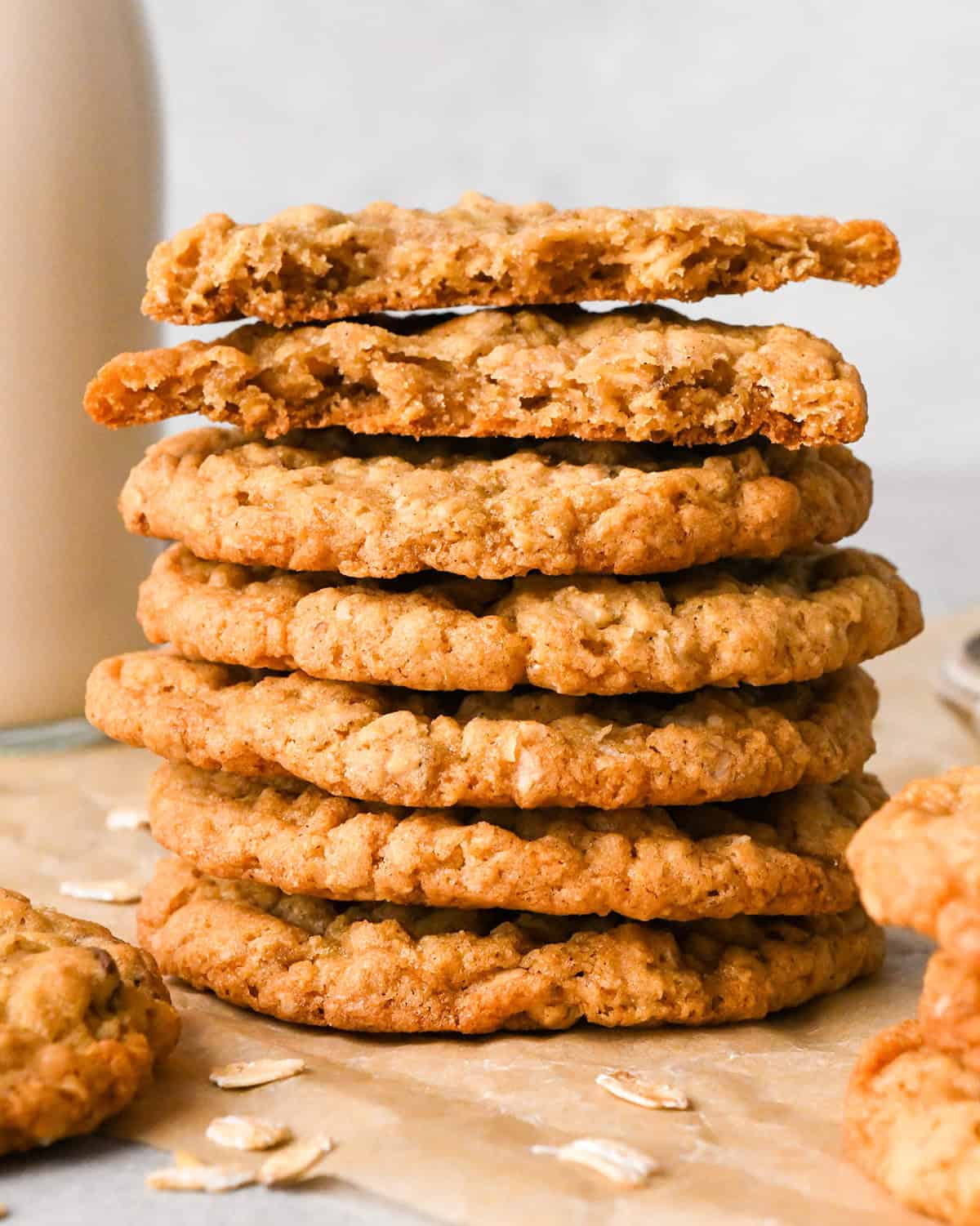 a stack of seven oatmeal cookies with the top cookie broken in half, placed on parchment paper with scattered  oats on the paper