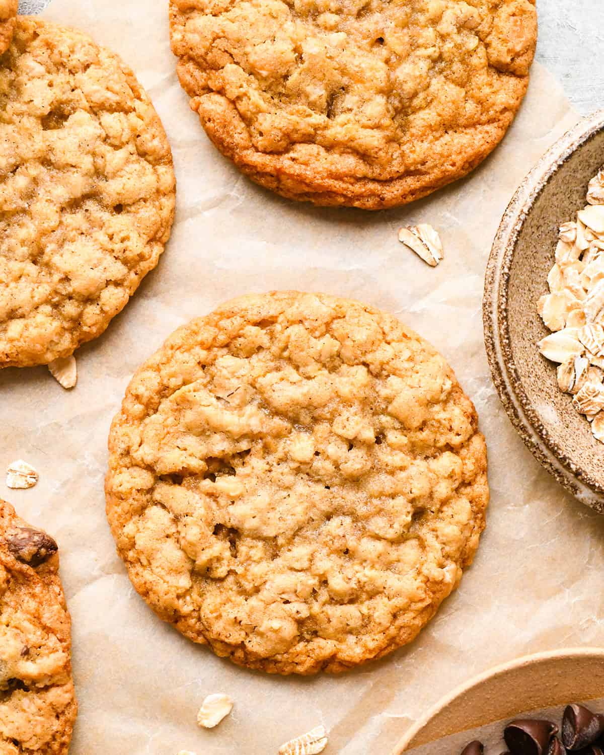 four oatmeal cookies on parchment paper next to a bowl of raw oats and a bowl of chocolate chips