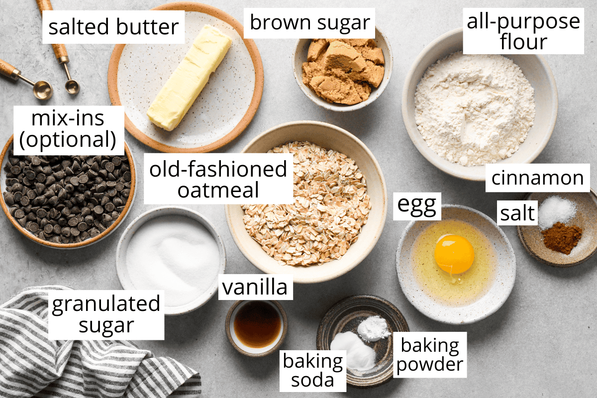 labeled ingredients for oatmeal cookies arranged on a surface, including butter, brown sugar, flour, oatmeal, egg, vanilla, sugar, baking soda, baking powder, cinnamon, salt, and optional mix-ins.