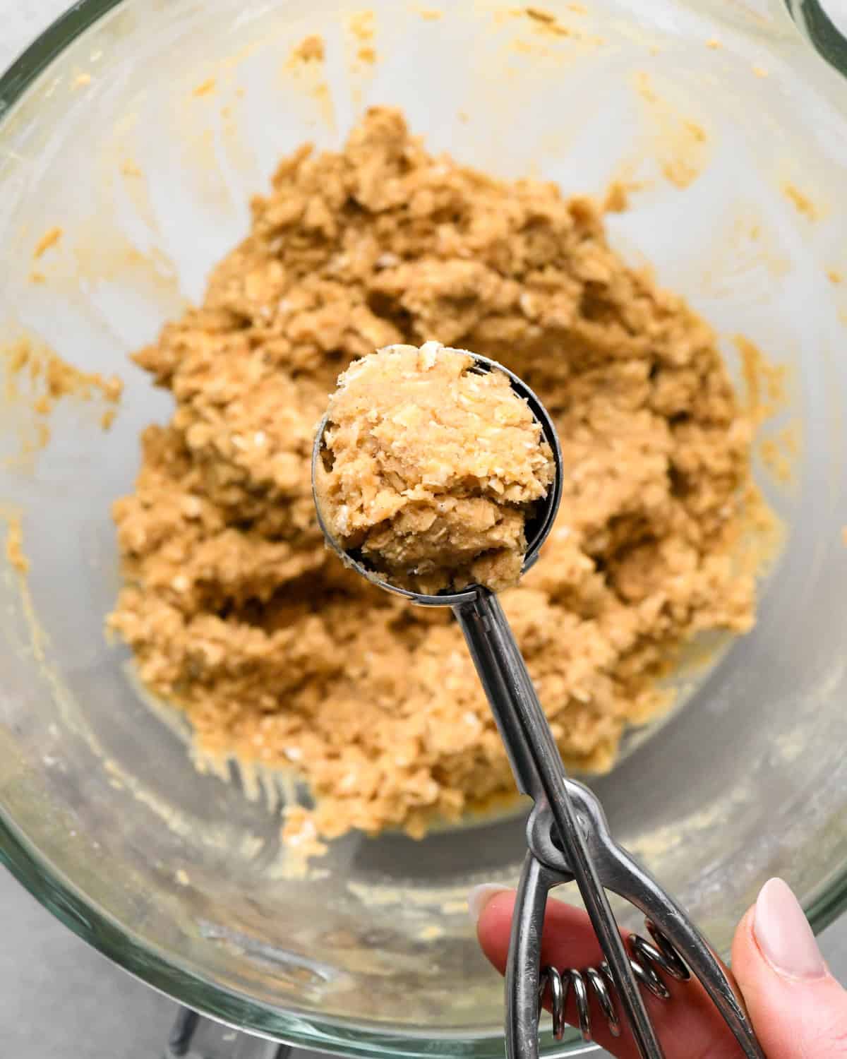 Cookie dough scoop being held over a glass mixing bowl filled with more dough making oatmeal cookies