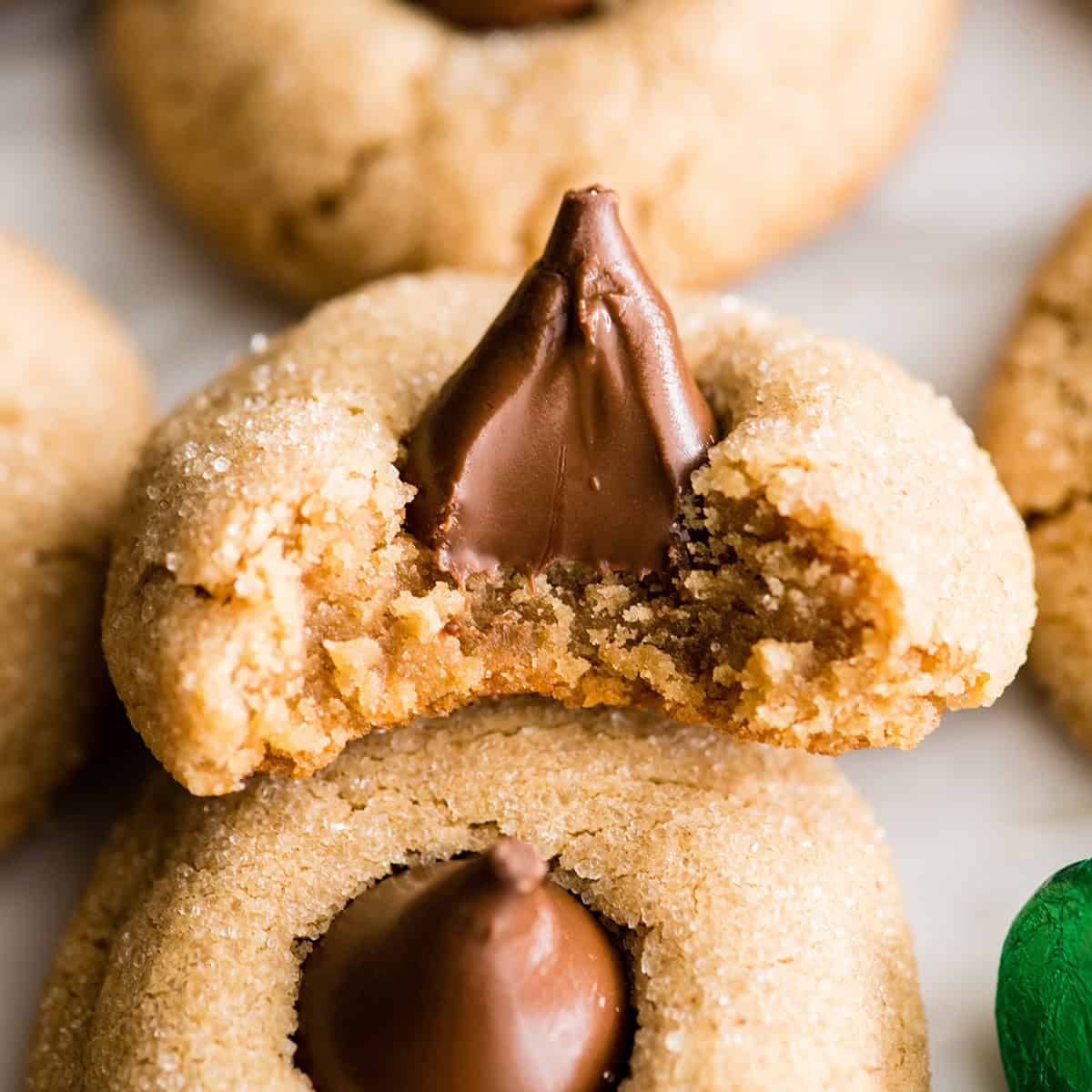 5 peanut butter blossom cookies, one has a bite taken out of it so they chewy texture and melty chocolate kiss are showing. There's a wrapped green kiss candy in the bottom right corner