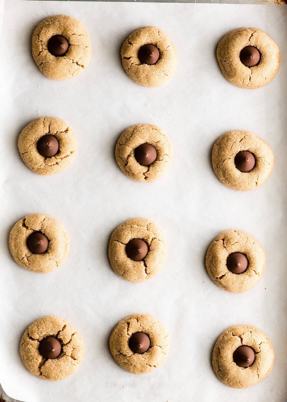making peanut butter blossoms - 12 peanut butter blossom cookies with the chocolate kisses pressed onto the top on a baking sheet lined with white parchment paper. cookies are in 4 rows of 3.
