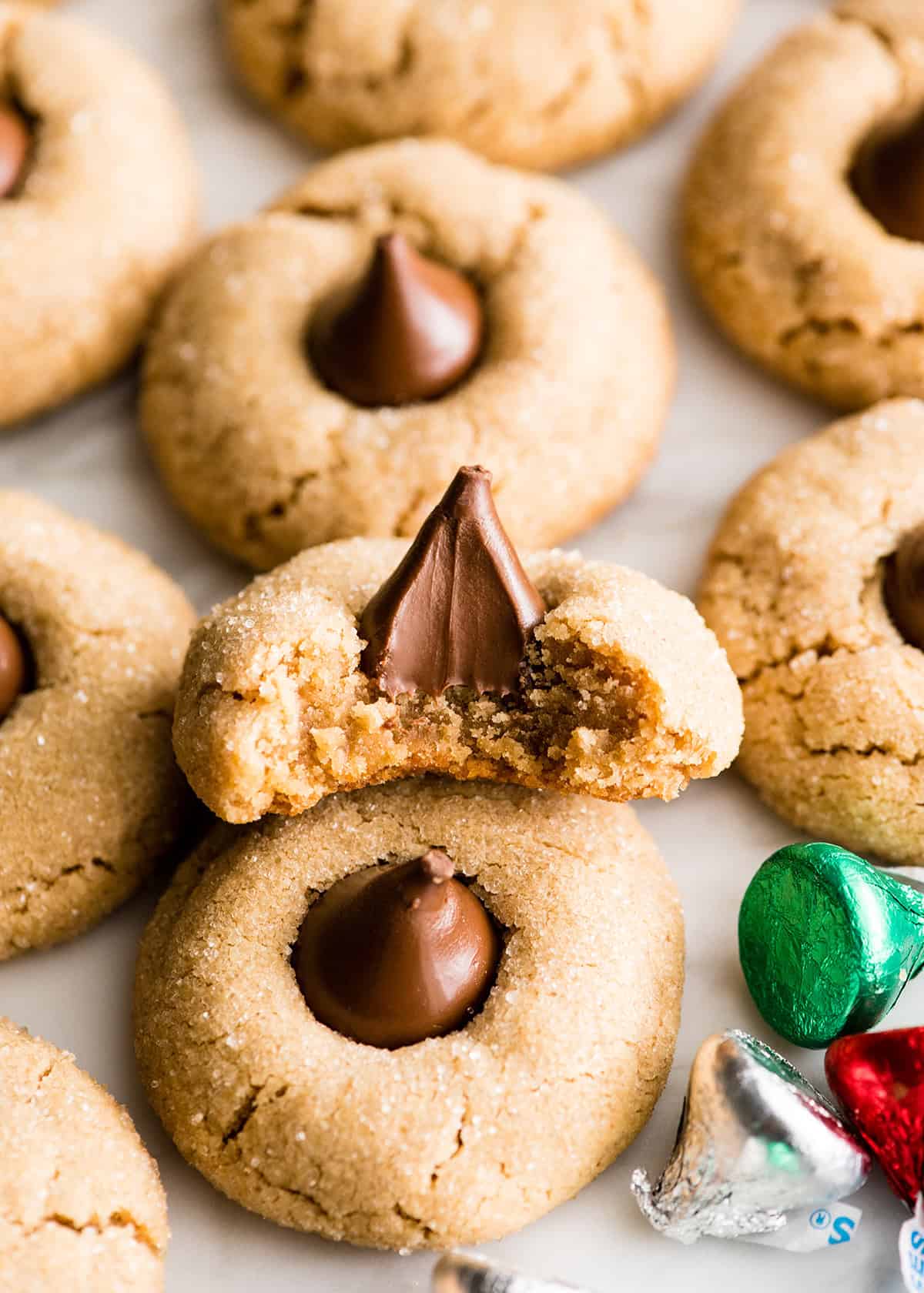8 peanut butter blossom cookies, one has a bite taken out of it so they chewy texture and melty chocolate kiss are showing. There's a wrapped green kiss candy in the bottom right corner