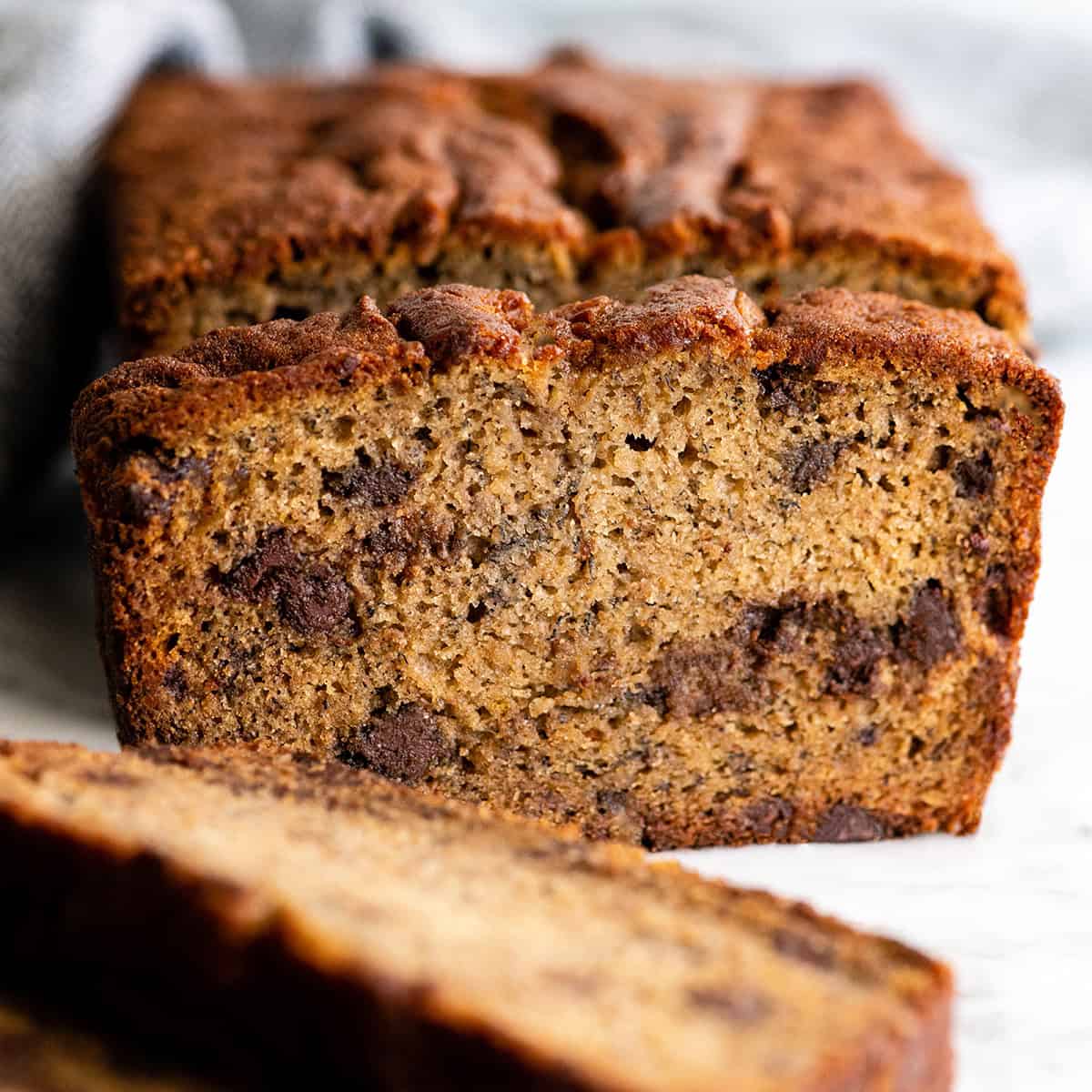 front view of a loaf of chocolate chip banana bread with slices cut out of it, some laying on the table one standing up in front of the loaf